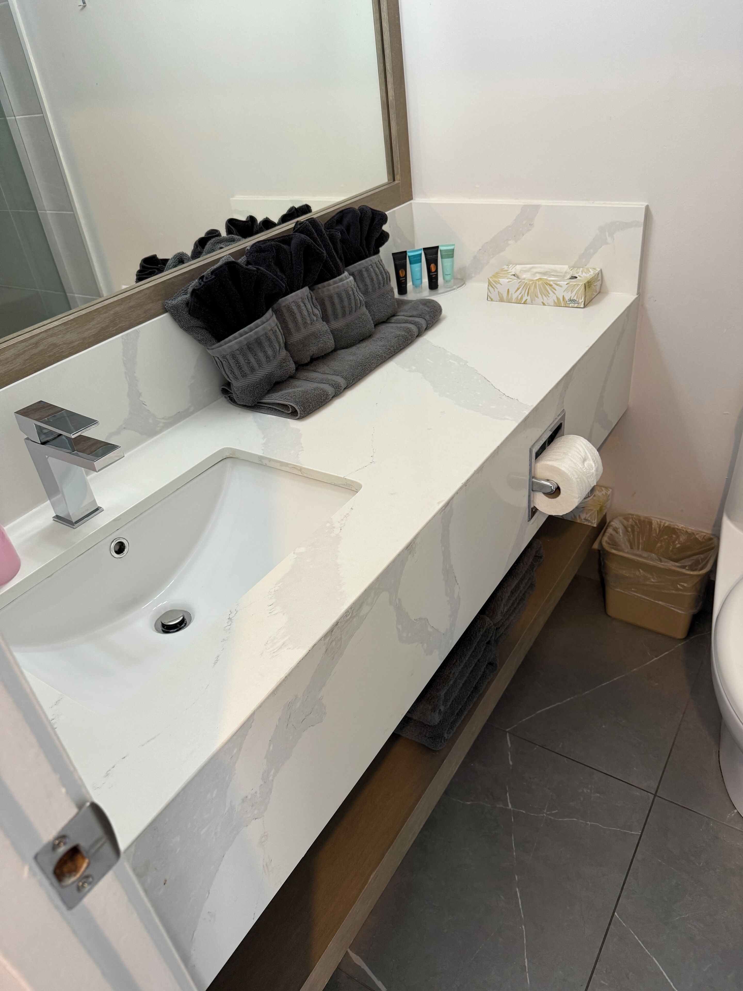 Un baño con lavamanos blanco, un espejo, toallero con toallas, artículos de tocador y un cubo de basura.