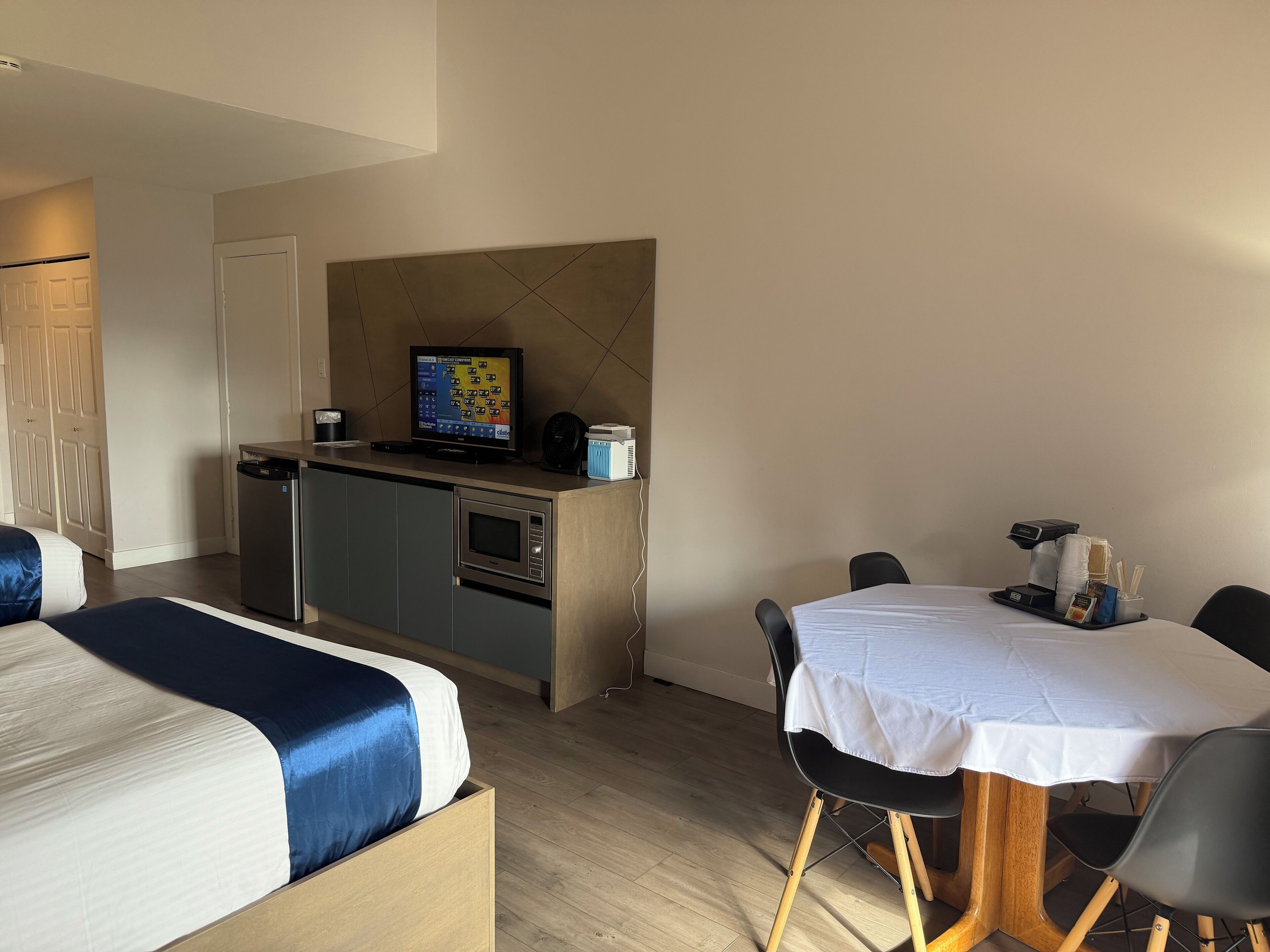 A hotel room with a bed, a small kitchenette, a dining table with chairs, and a television.