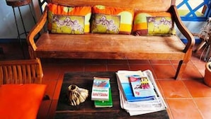 Ein Holzsofa mit bunten Kissen, ein Holzstuhl und ein Tisch mit Büchern und einer Pflanze.