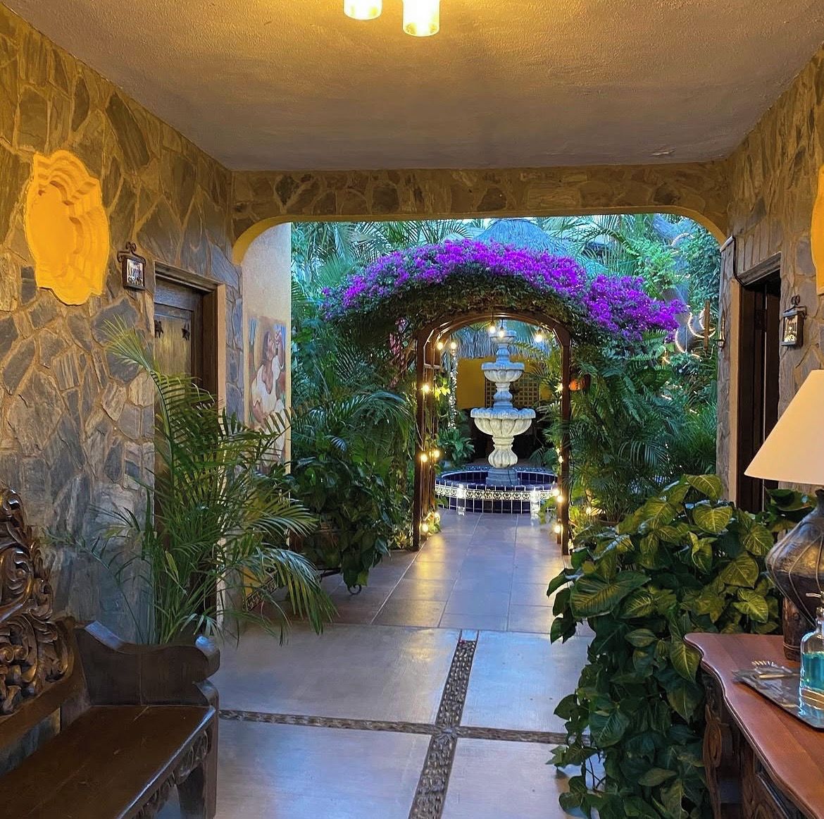 A hallway with a fountain, stone walls, and lush greenery.