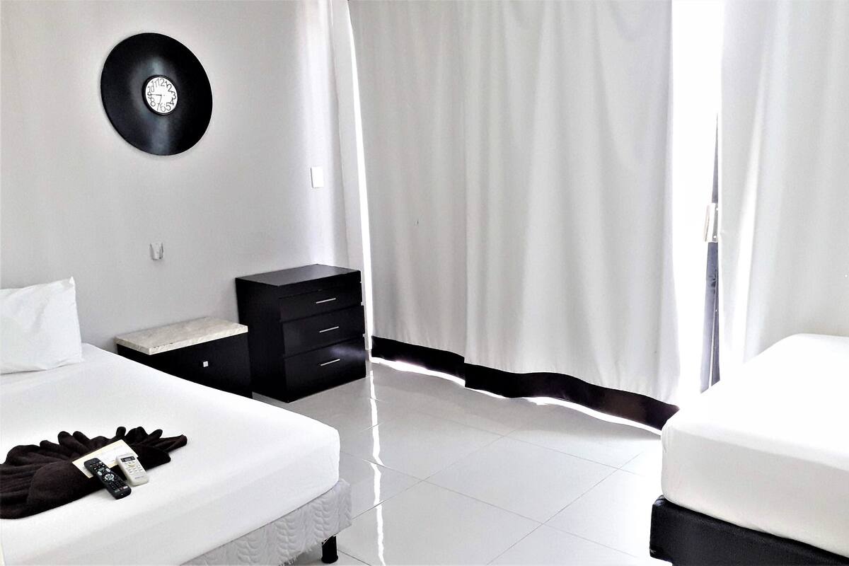 A bedroom with a bed, a bedside table, a black clock on the wall, and a curtain.