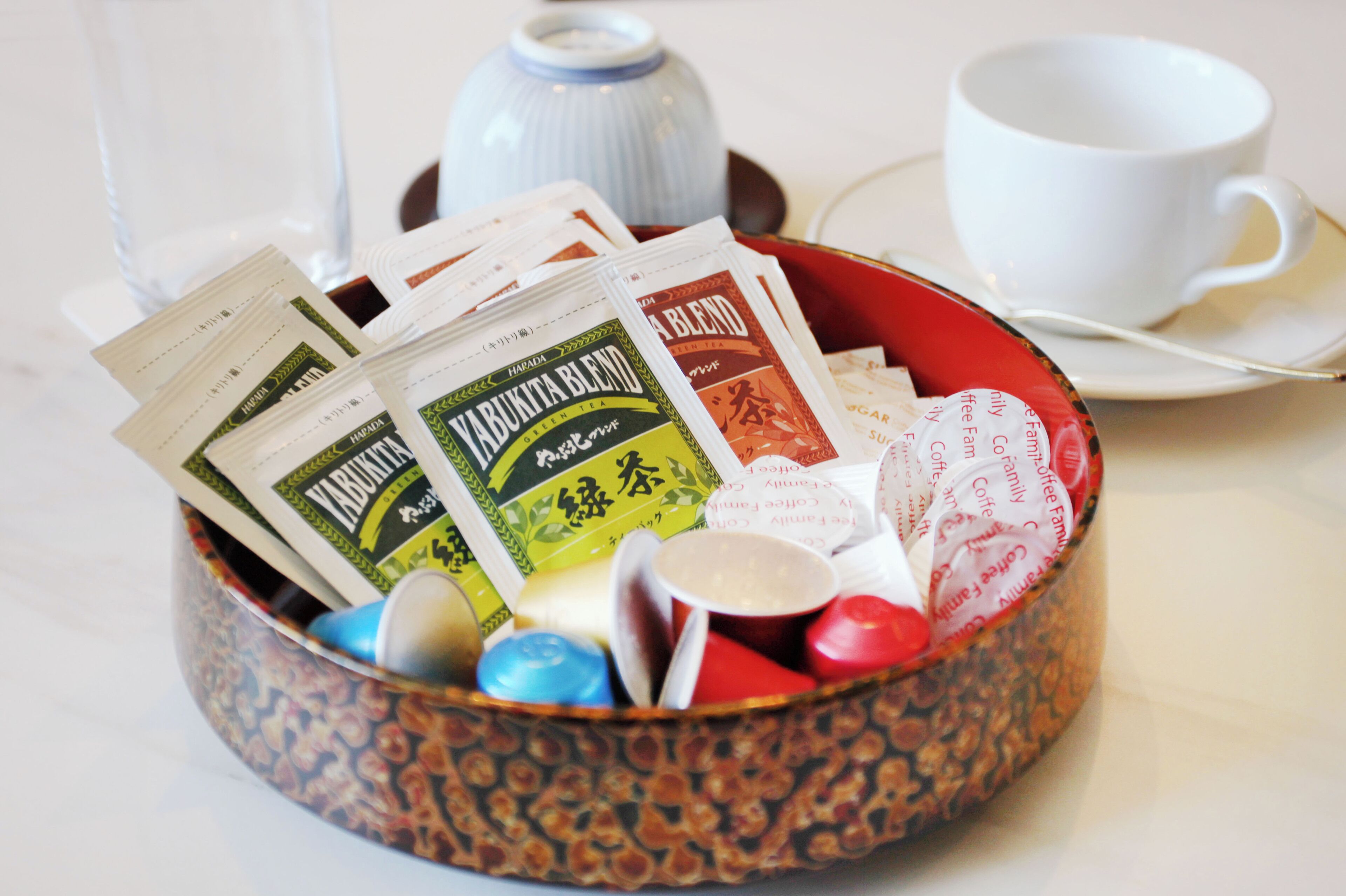 A round, patterned bowl filled with coffee packets and a white cup on a saucer.