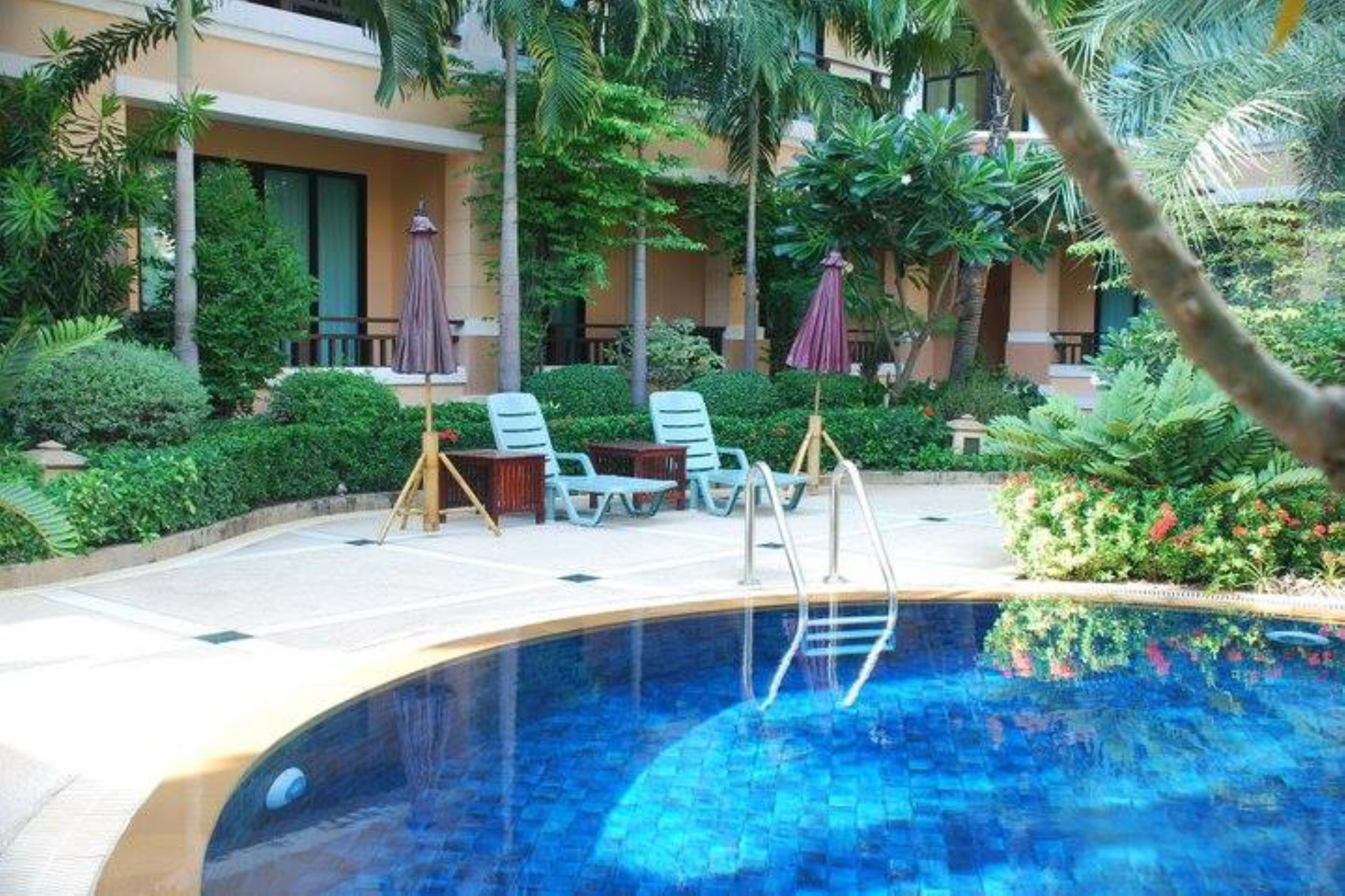 A swimming pool with lounge chairs and umbrellas, surrounded by lush greenery and a building in the background.