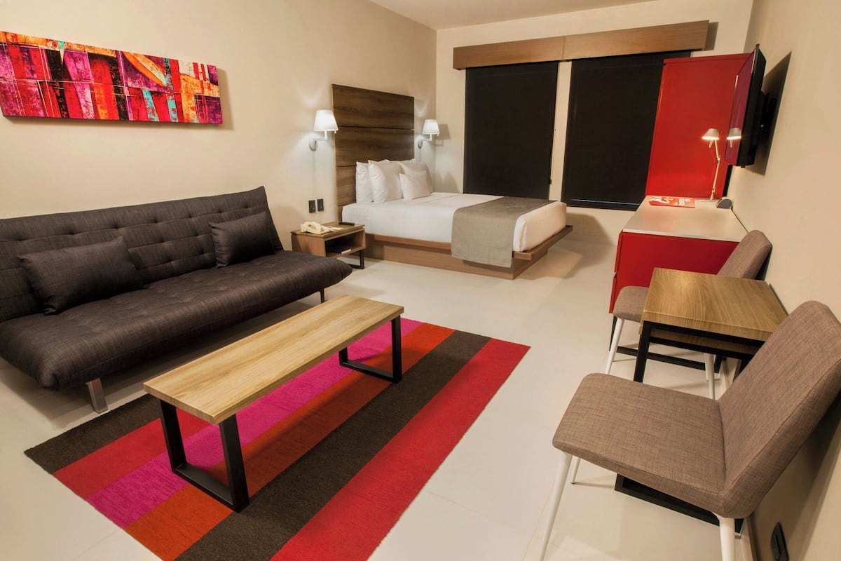 A hotel room with a bed, a sofa, a table, a chair, a TV, and a painting on the wall.