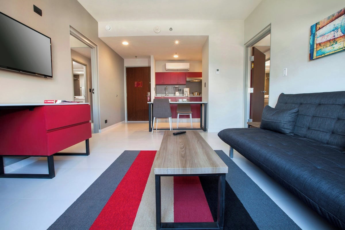 A modern living room with a red sofa, a grey couch, and a wooden coffee table. There is a kitchen in the background with red cabinets and a white countertop.