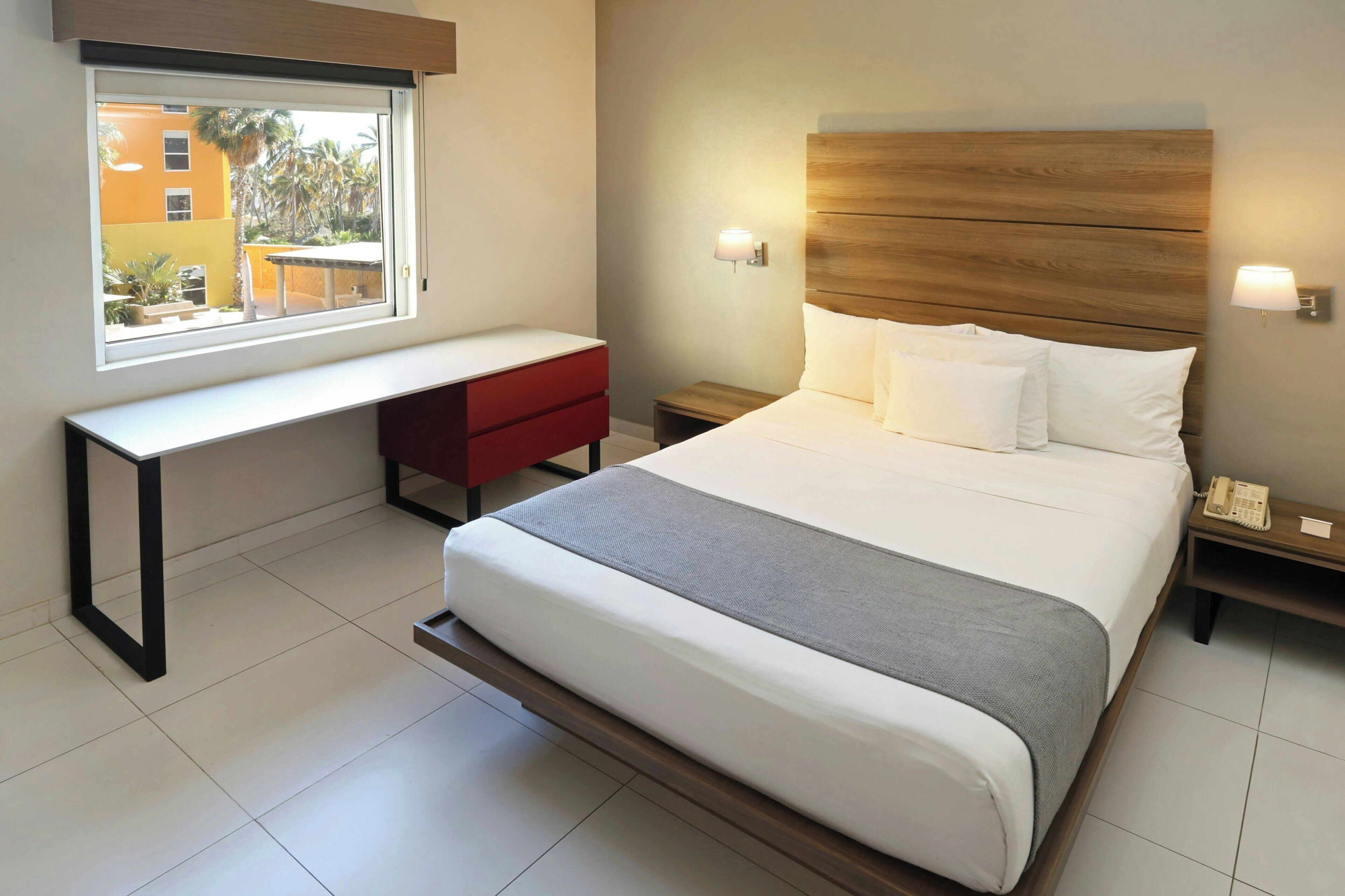 A hotel room with a bed, bedside tables, a desk, and a window with a view of buildings.