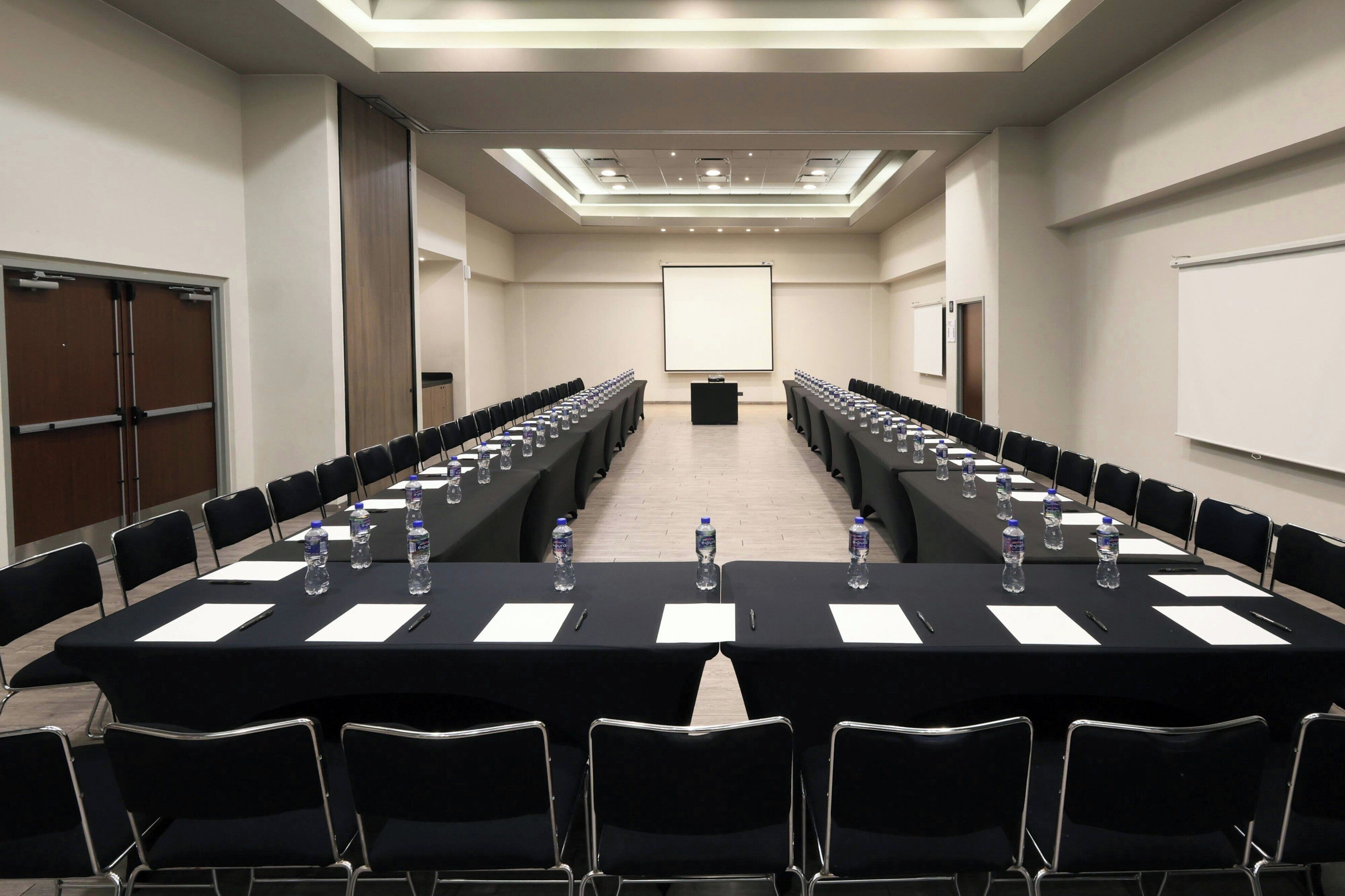 A conference room with long tables, chairs, water bottles, and a projector screen.
