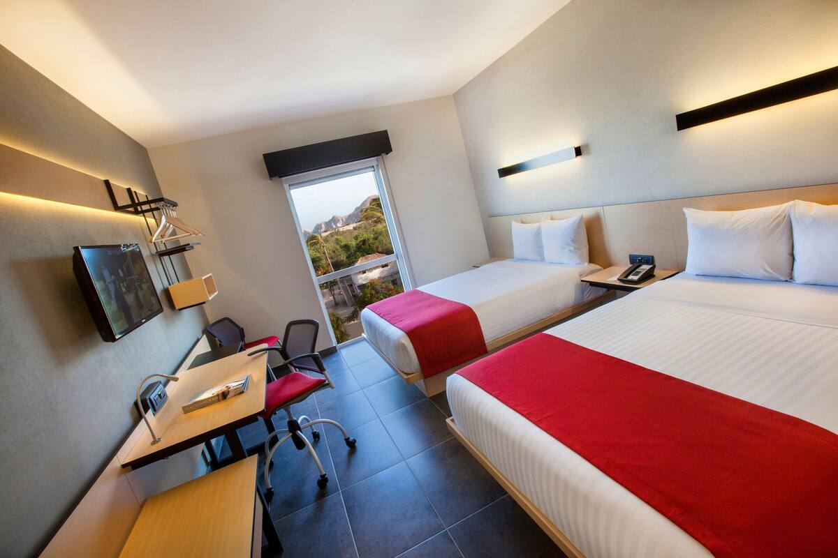 A hotel room with two beds, a desk with a chair, a TV, and a window with a view of the outdoors.