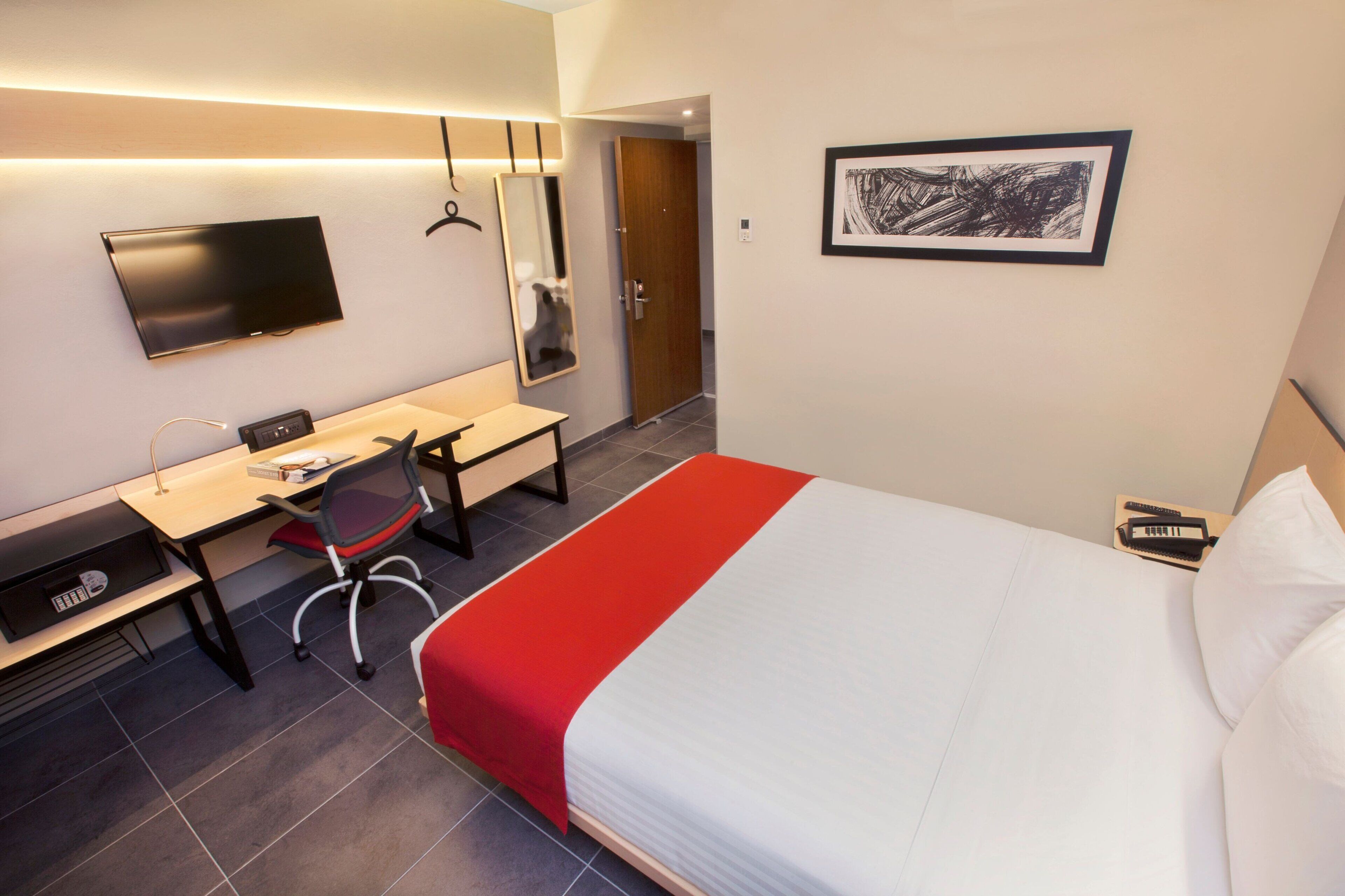 A hotel room with a bed, a desk, a chair, a TV, and a door.