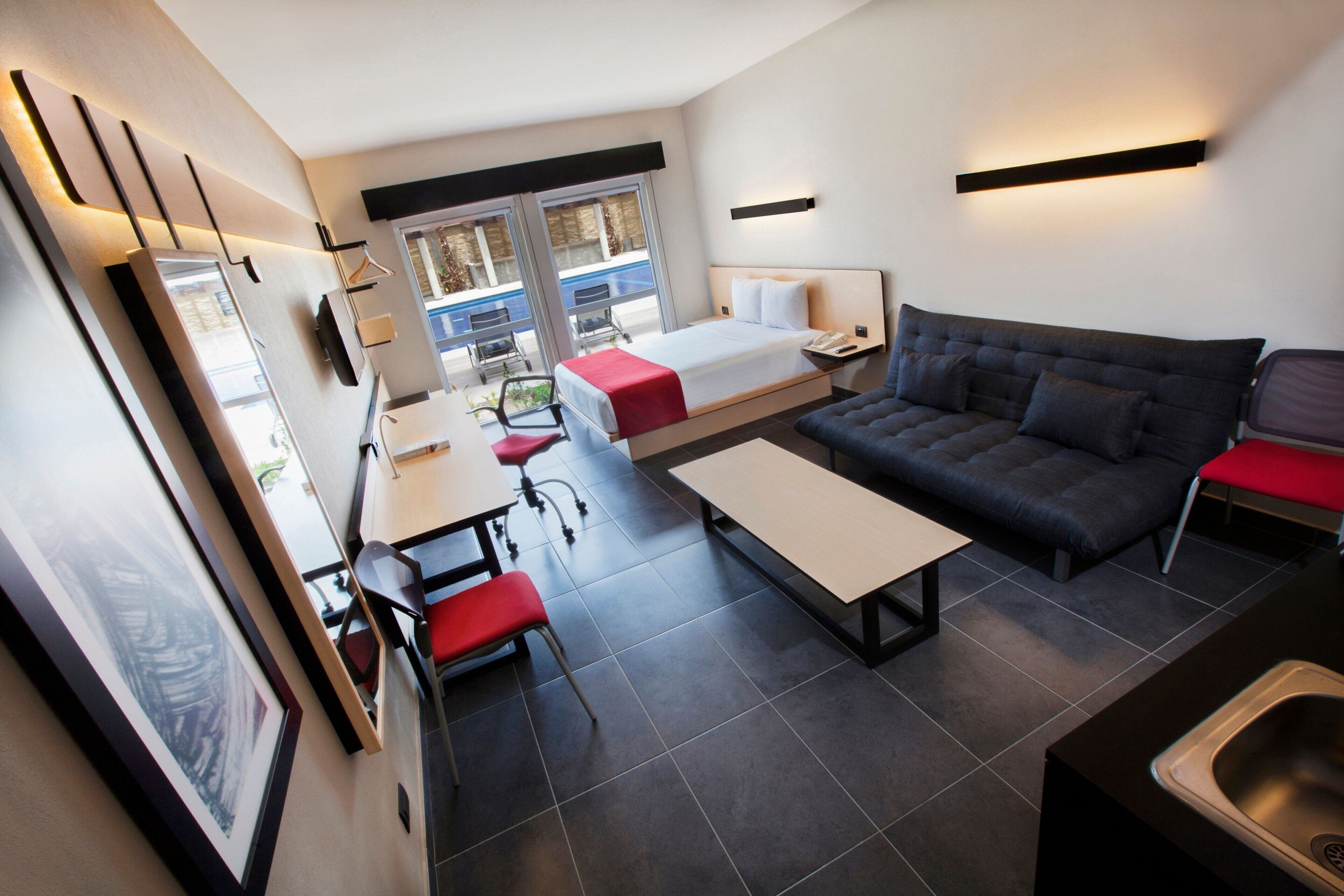 Quarto de hotel moderno com cama, sofá, mesa de jantar e uma pequena cozinha.