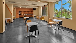 Un'area pranzo moderna con tavoli e sedie, una cucina sullo sfondo e ampie finestre che si affacciano su palme.