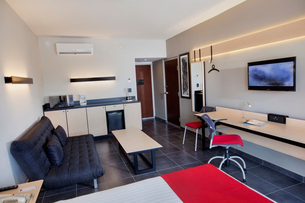 A modern hotel room with a sofa, a dining table, a TV, and a kitchenette.
