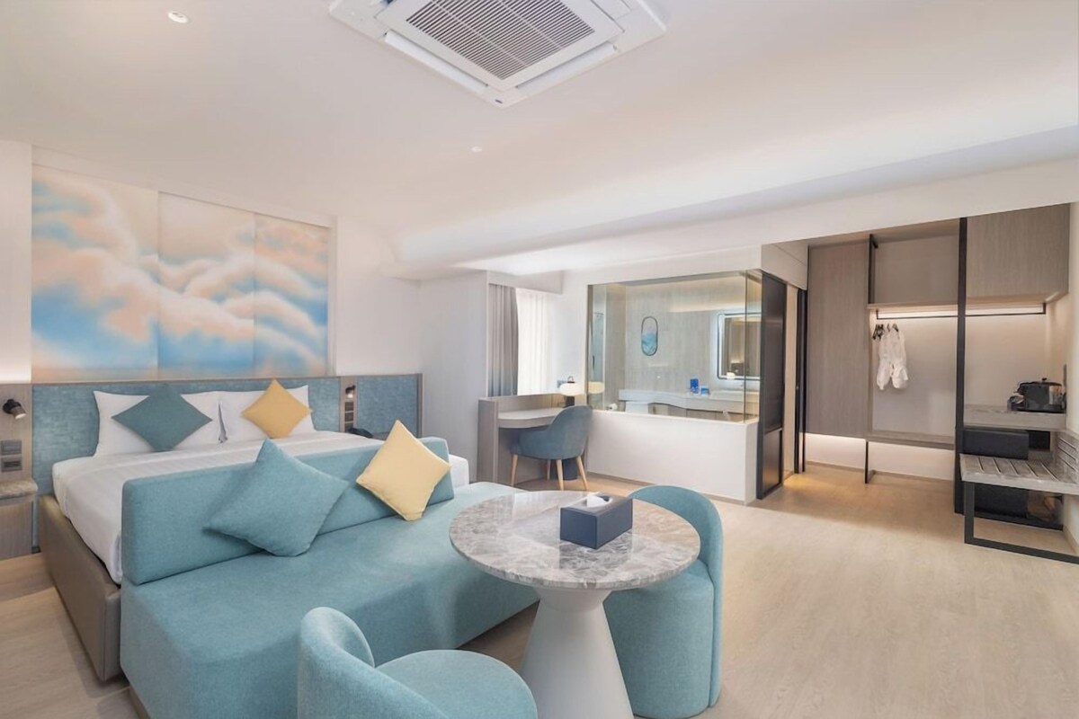 A modern hotel room with a bed, a seating area, a desk, and a walk-in closet.