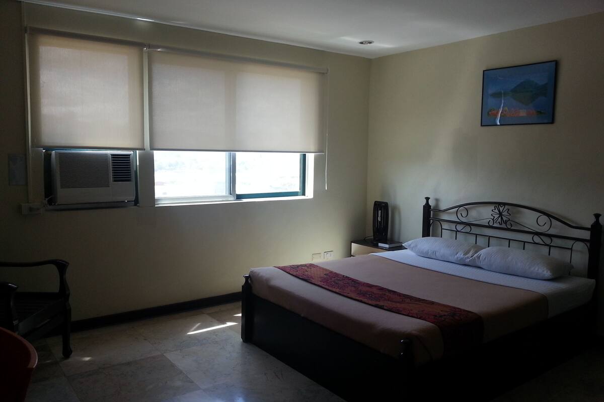 A bedroom with a bed, a nightstand, a chair, a window with blinds, and an air conditioner.