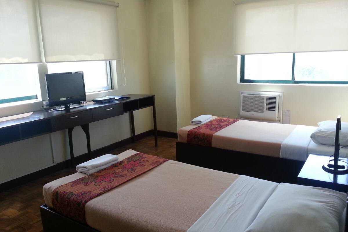 A hotel room with two beds, a desk, a television, and an air conditioning unit.