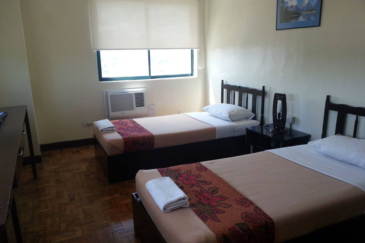 A hotel room with two beds, a window with blinds, a wall-mounted air conditioner, a bedside table with a lamp, and a painting on the wall.