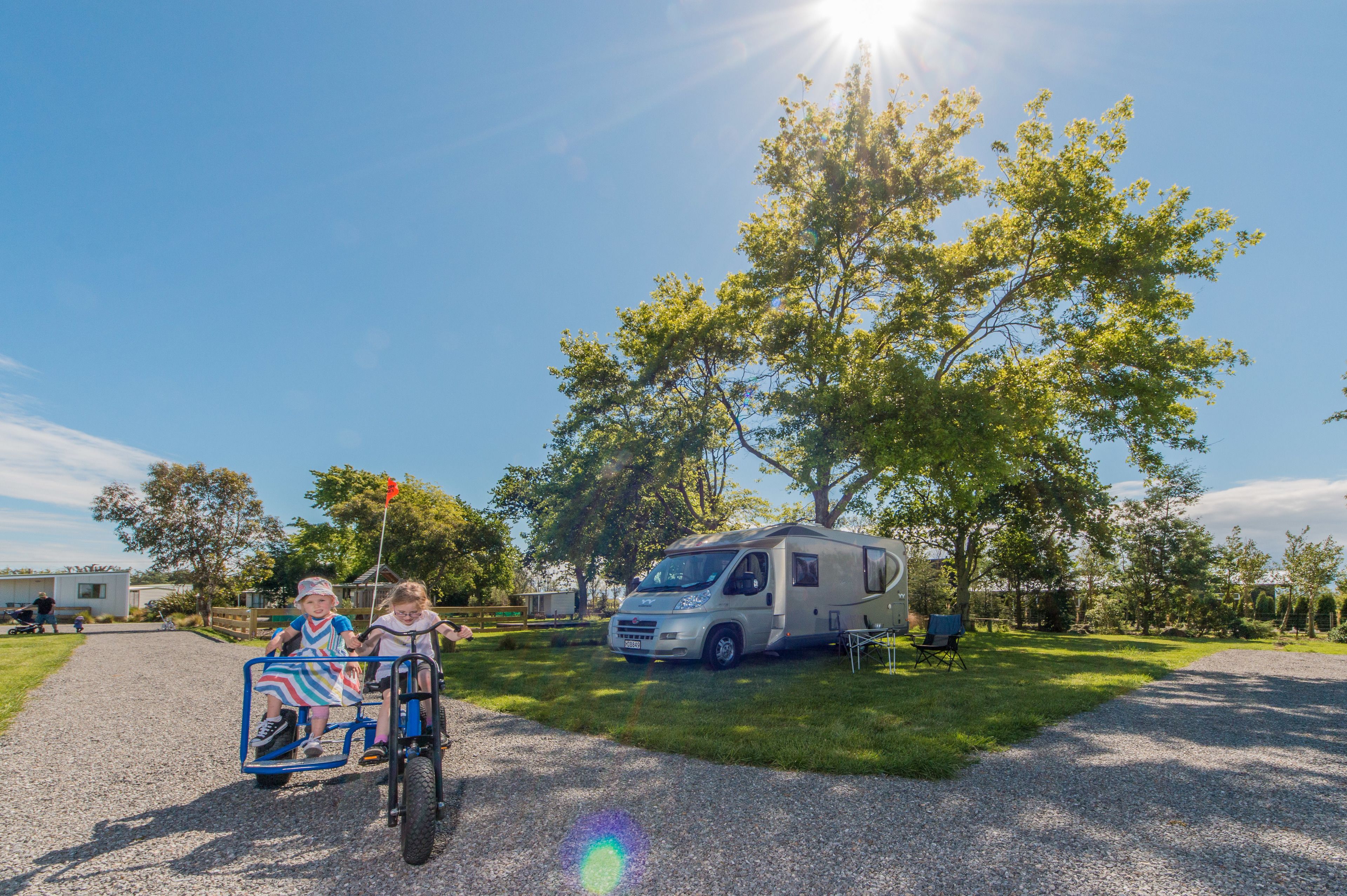 A gravel driveway with a white RV parked on the grass, a large tree, and two children on a tricycle.