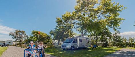A gravel driveway with a white RV parked on the grass, a large tree, and two children on a tricycle.
