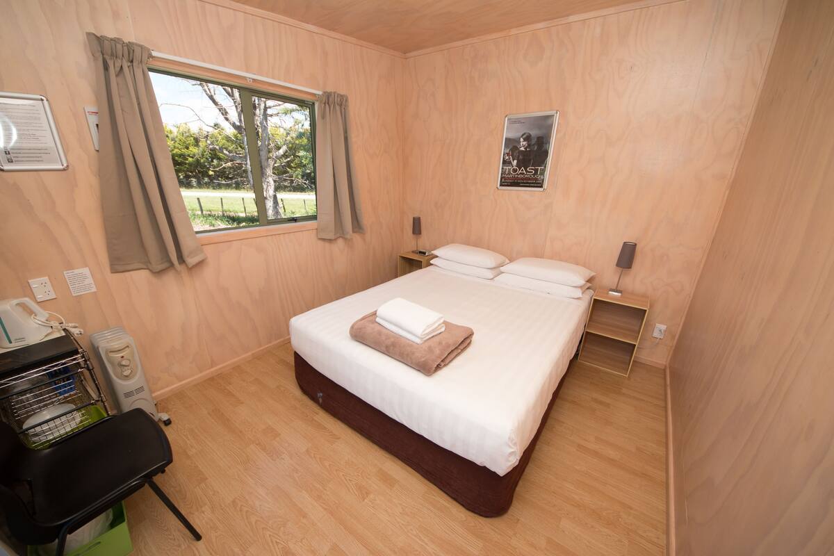 Ein Zimmer mit Holzboden, einem Bett mit weißer Bettwäsche und einem braunen Handtuch, einem Fenster mit Vorhängen, einem Poster an der Wand und einem kleinen Nachttisch.