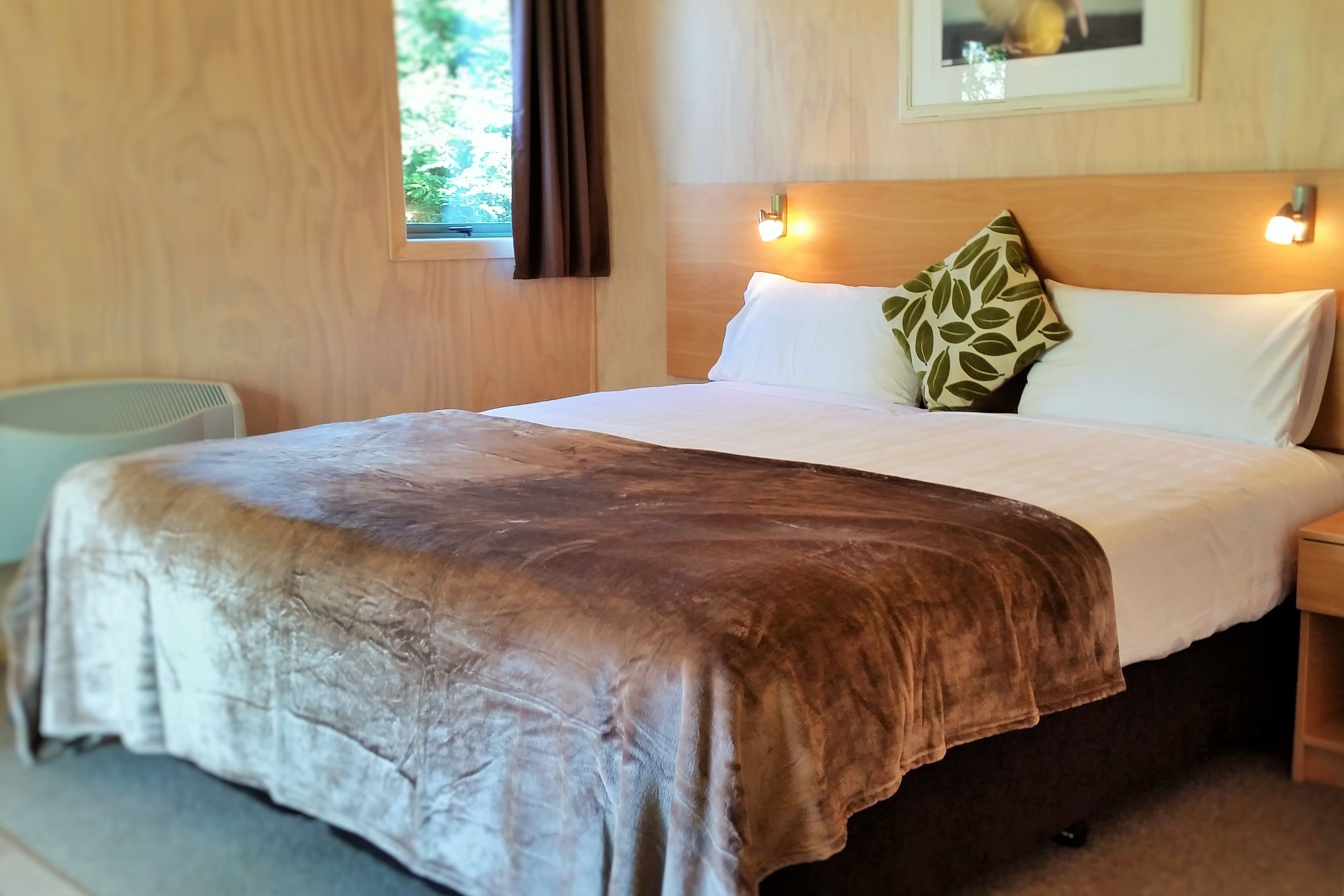 A hotel room with a wooden headboard, a bed with a brown blanket, white pillows, and a green leaf-patterned pillow.