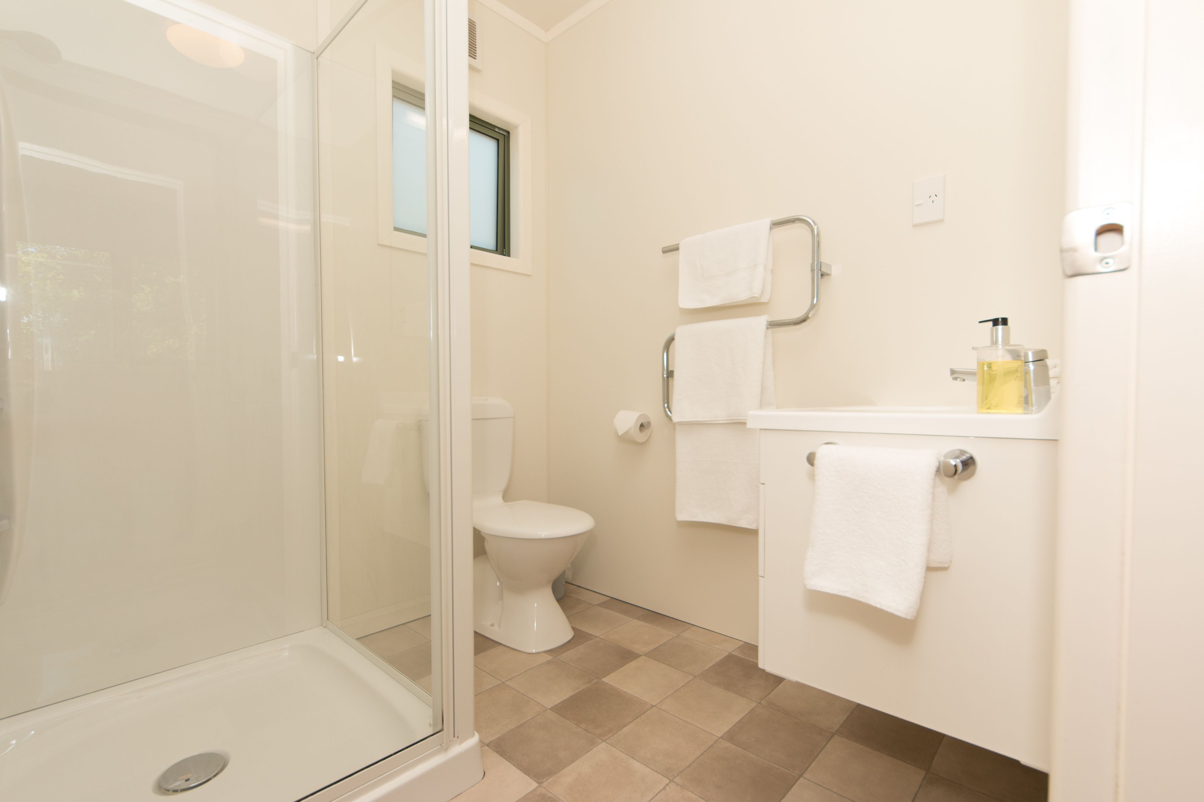 A bathroom with a glass shower enclosure, a toilet, a sink with a soap dispenser, and two towels hanging on a rack.