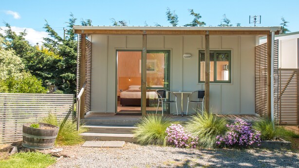 Una cabaña pequeña de una sola habitación con una cama, una mesa y sillas afuera.