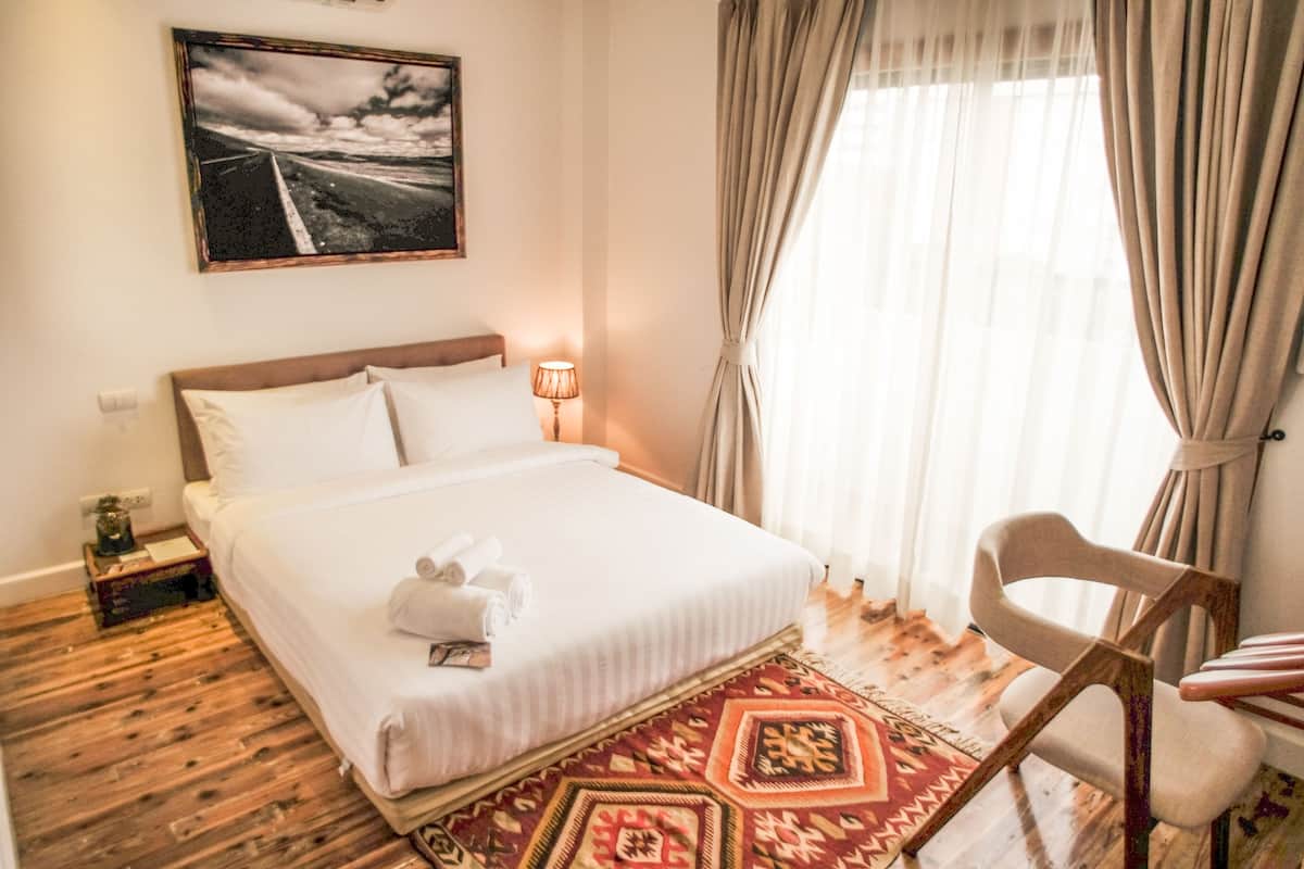 A hotel room with a large bed, a wooden floor, a patterned rug, a chair, a lamp, and a painting on the wall.