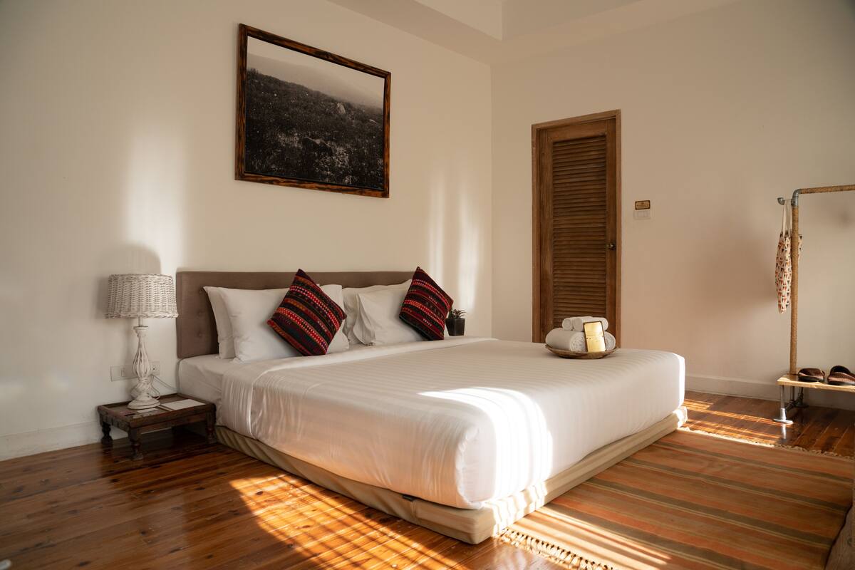 A bedroom with a large bed, a nightstand, a lamp, a framed picture on the wall, and a wooden door.