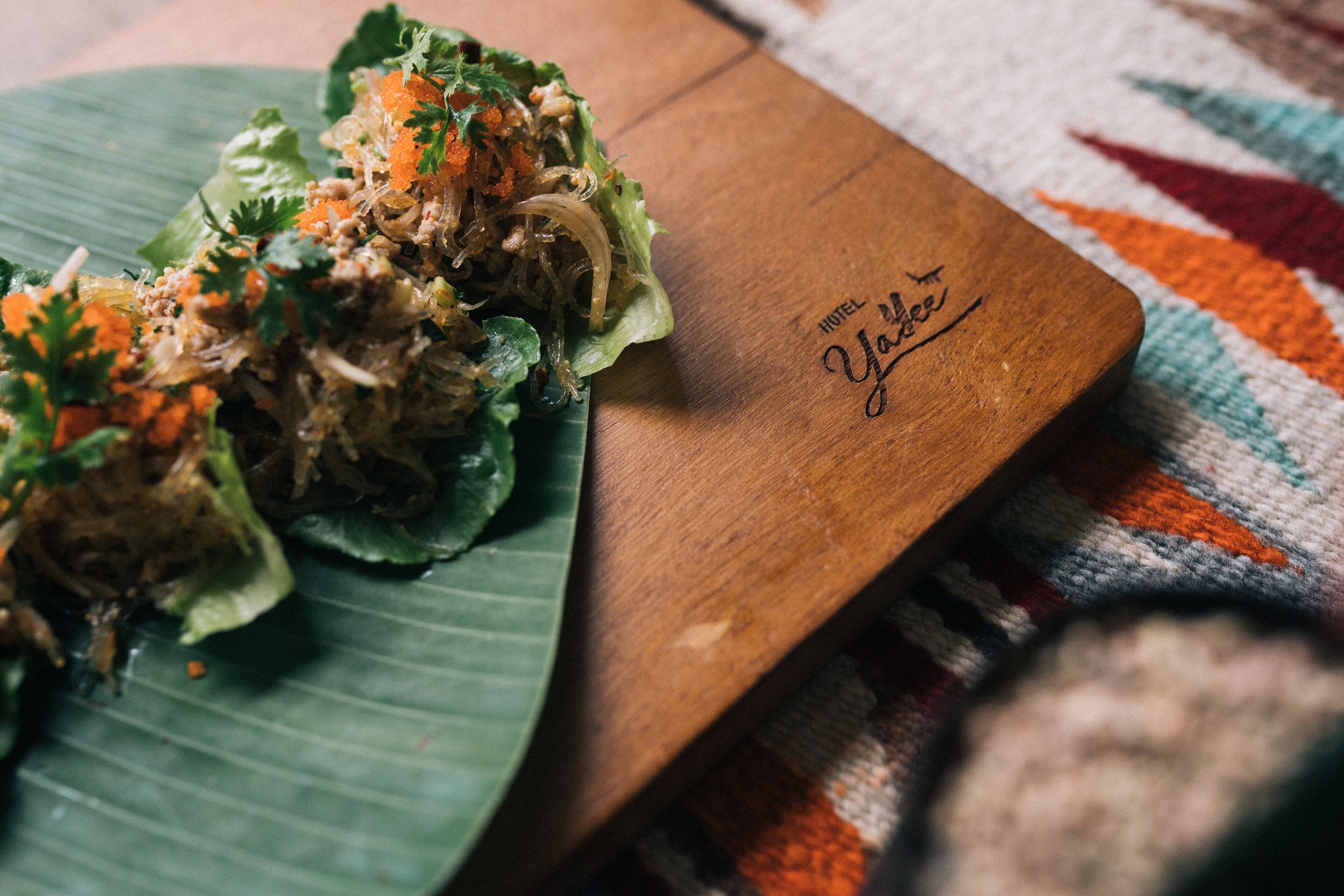 A wooden serving board with a salad on a green leaf, placed on a colorful woven mat.