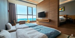 A hotel room with two beds, a TV, a desk, and a view of the sea.