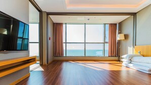 Camera d'albergo con un'ampia finestra, pavimento in legno, televisione a schermo piatto e un letto con lenzuola bianche.