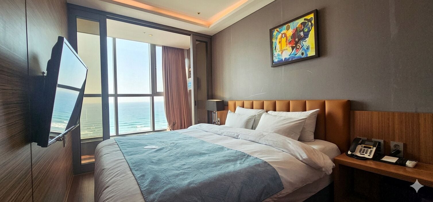 Une chambre d’hôtel avec un lit, un téléphone sur la table de chevet, un tableau au mur et une fenêtre donnant sur l’océan.