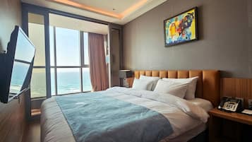 A hotel room with a bed, a bedside telephone, a painting on the wall, and a window with a view of the ocean.