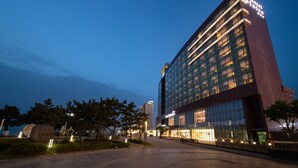 Un hotel moderno con una facciata in vetro ben visibile, un ingresso luminoso e un'area verde con alberi e un vialetto.