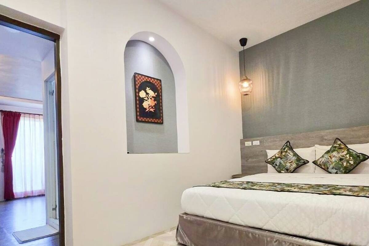 Quarto com cama, mesa de cabeceira, luminária na parede e porta que dá para outro cômodo.