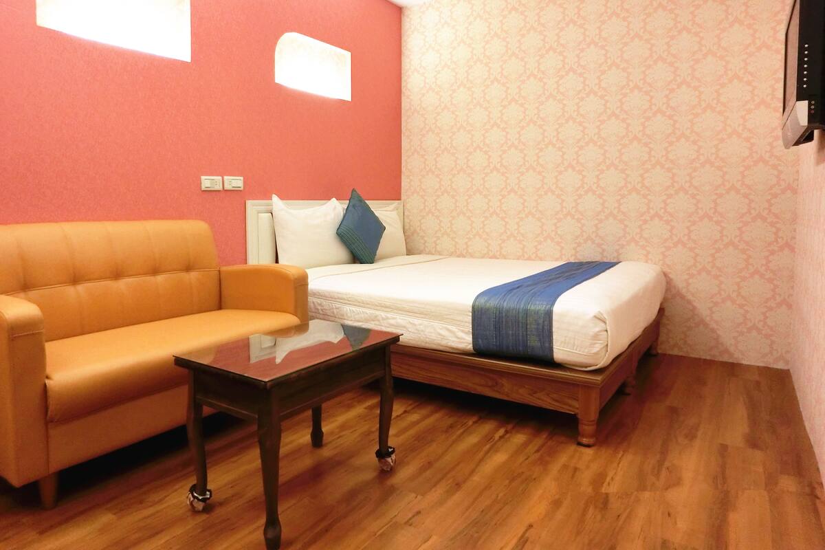 Quarto com piso de madeira, uma cama com cobertor azul, um sofá e uma mesa pequena.