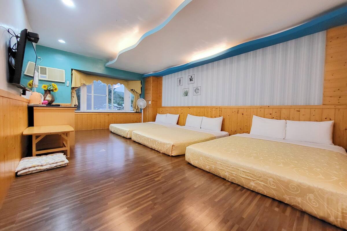 Quarto de hotel com duas camas, uma escrivaninha, uma cadeira, uma janela com cortinas e uma parede com quadros.