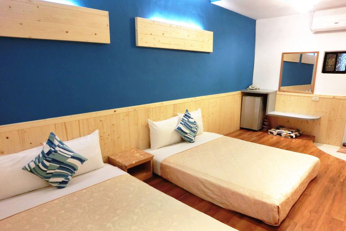 Quarto de hotel com duas camas, cabeceiras de madeira, uma mesinha de cabeceira pequena de madeira, um espelho e um banco de madeira.