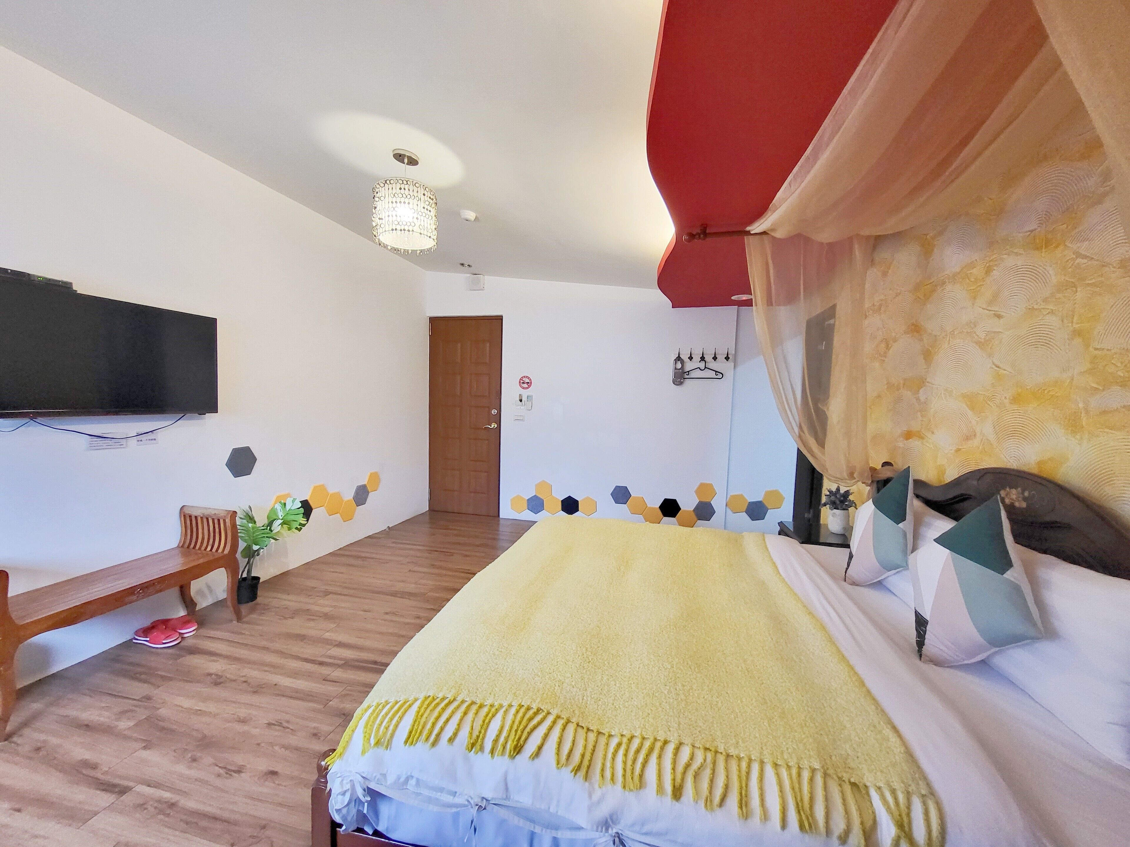 A bedroom with a bed, a TV mounted on the wall, a wooden bench, a door, and a wall with hexagonal patterns.