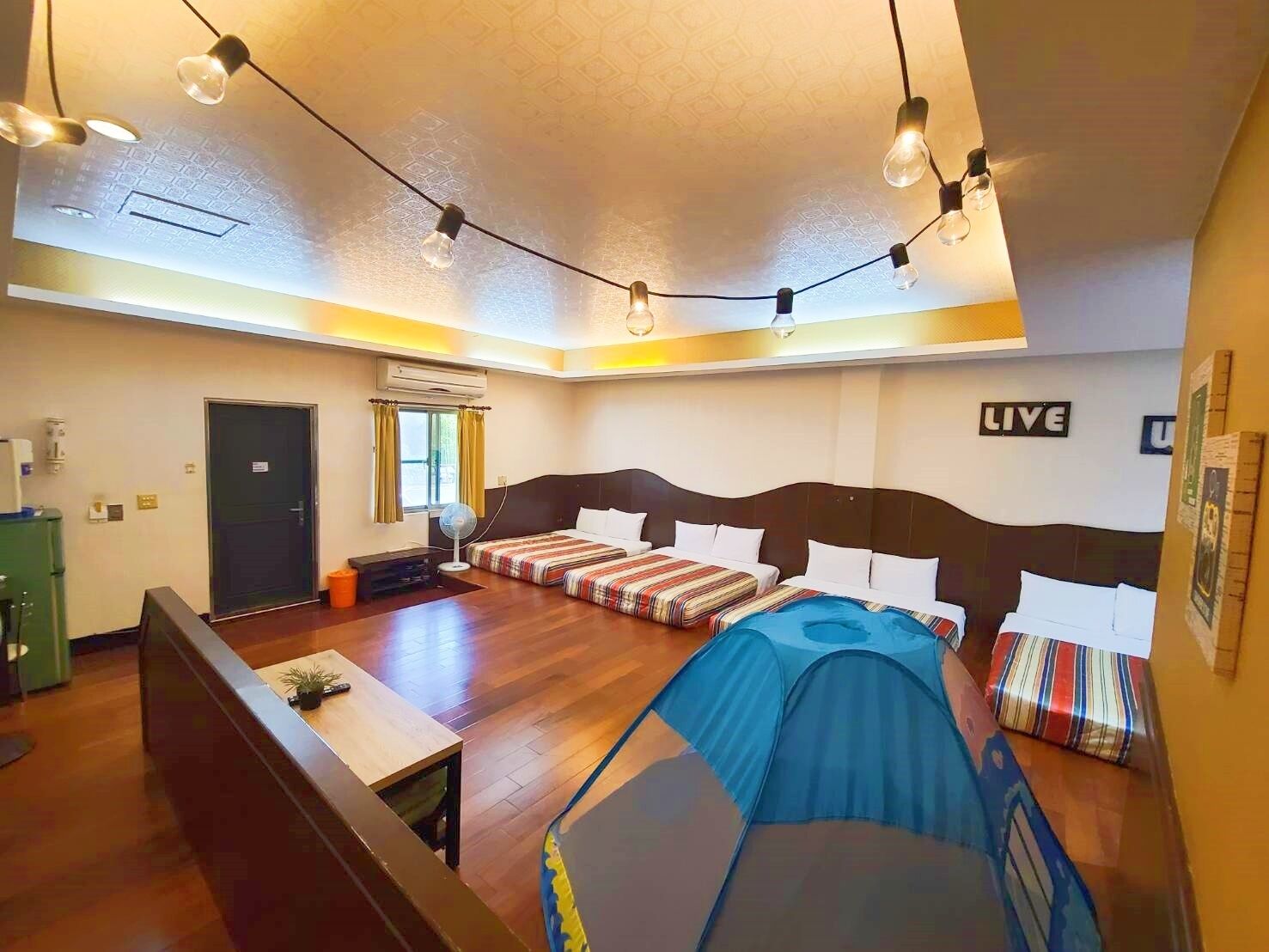 A hotel room with a large bed, a smaller bed, a desk, and a chair. There is a window with curtains, a door, and a wall-mounted air conditioner.