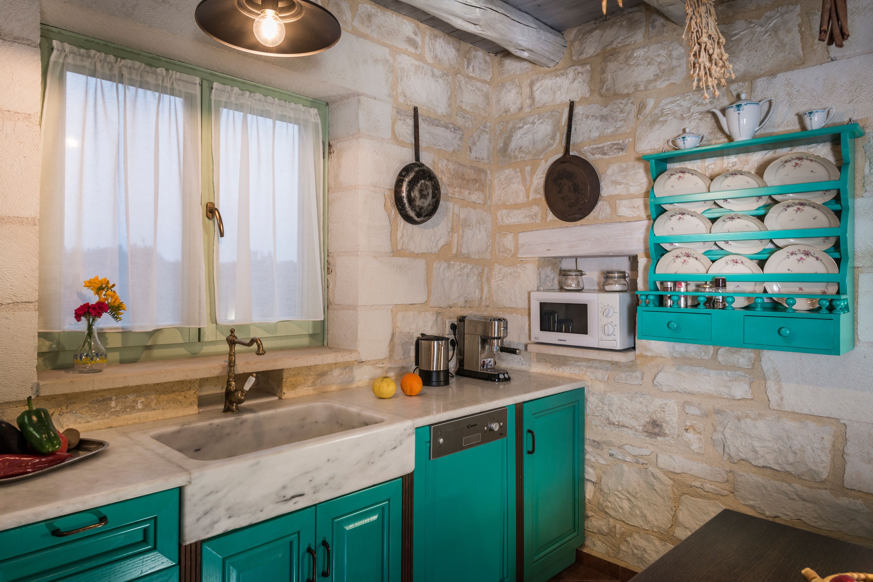 A kitchen with teal cabinets, a stone wall, a window with curtains, a coffee maker, and a dish rack.