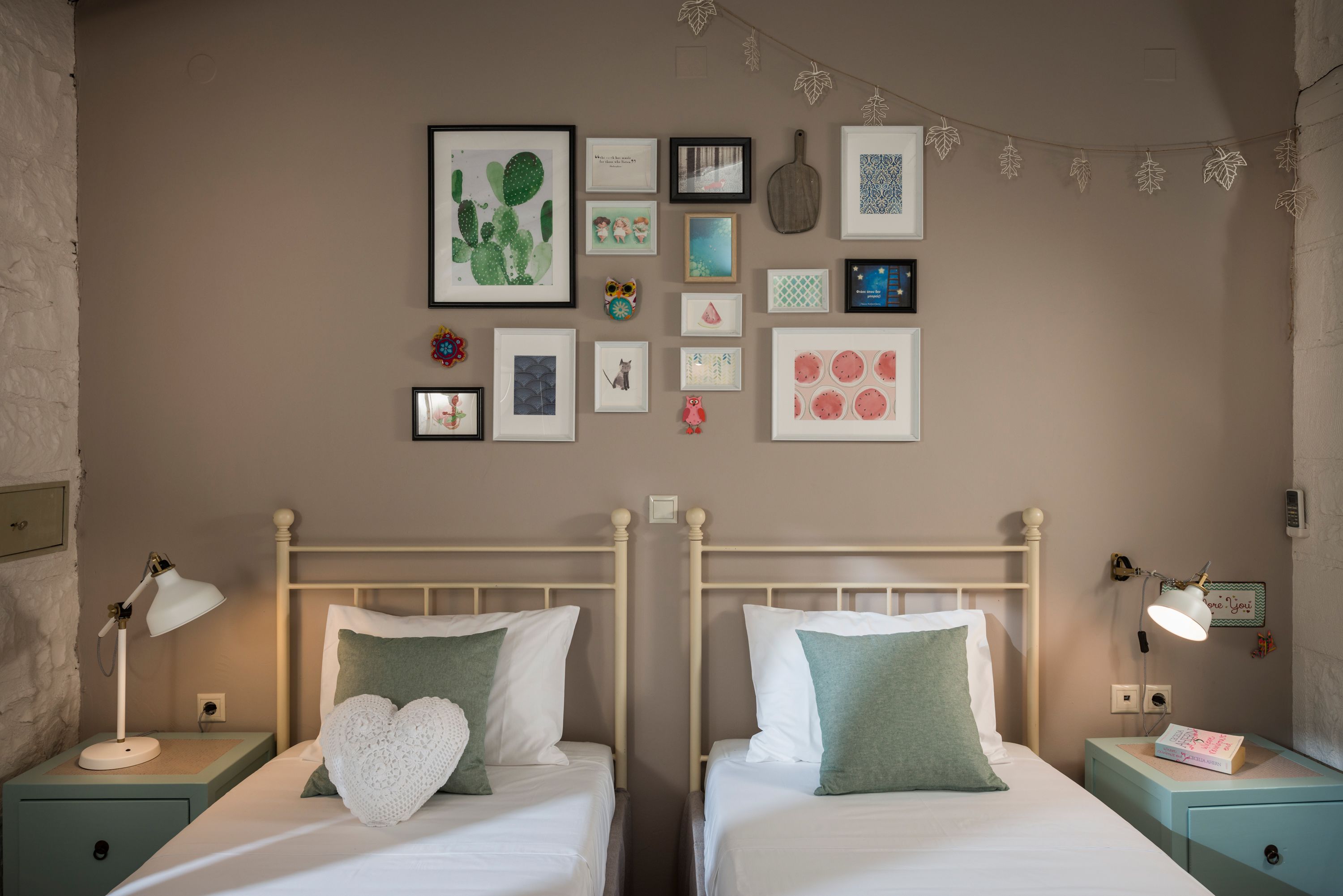 A room with two beds, each with a nightstand, a lamp, and a framed picture collection on the wall.