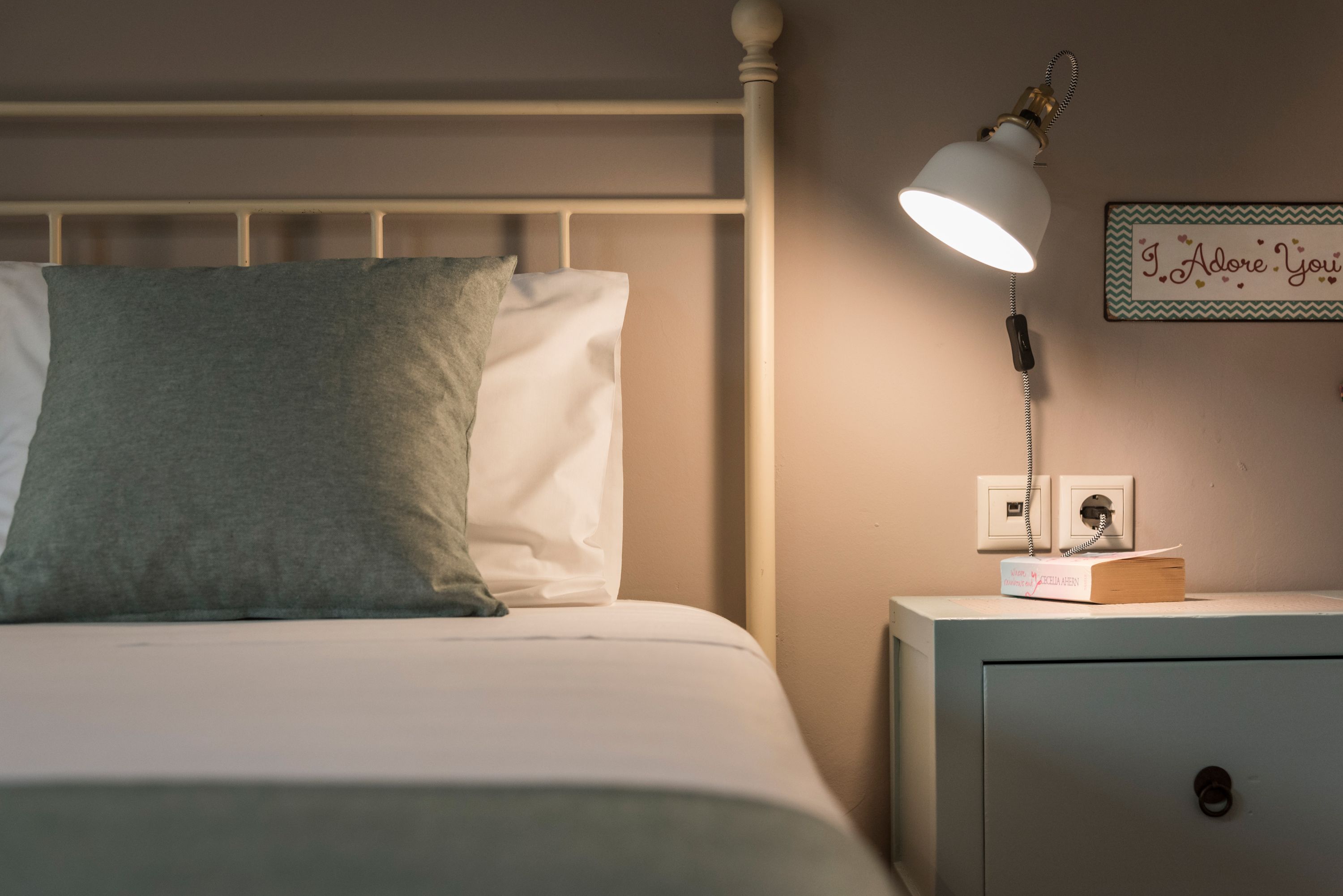 Un lit bien fait, avec un oreiller vert, une lampe de chevet, une petite table de chevet sur laquelle repose un livre, et un panneau fixé au mur portant l'inscription : « Je t'adore ».