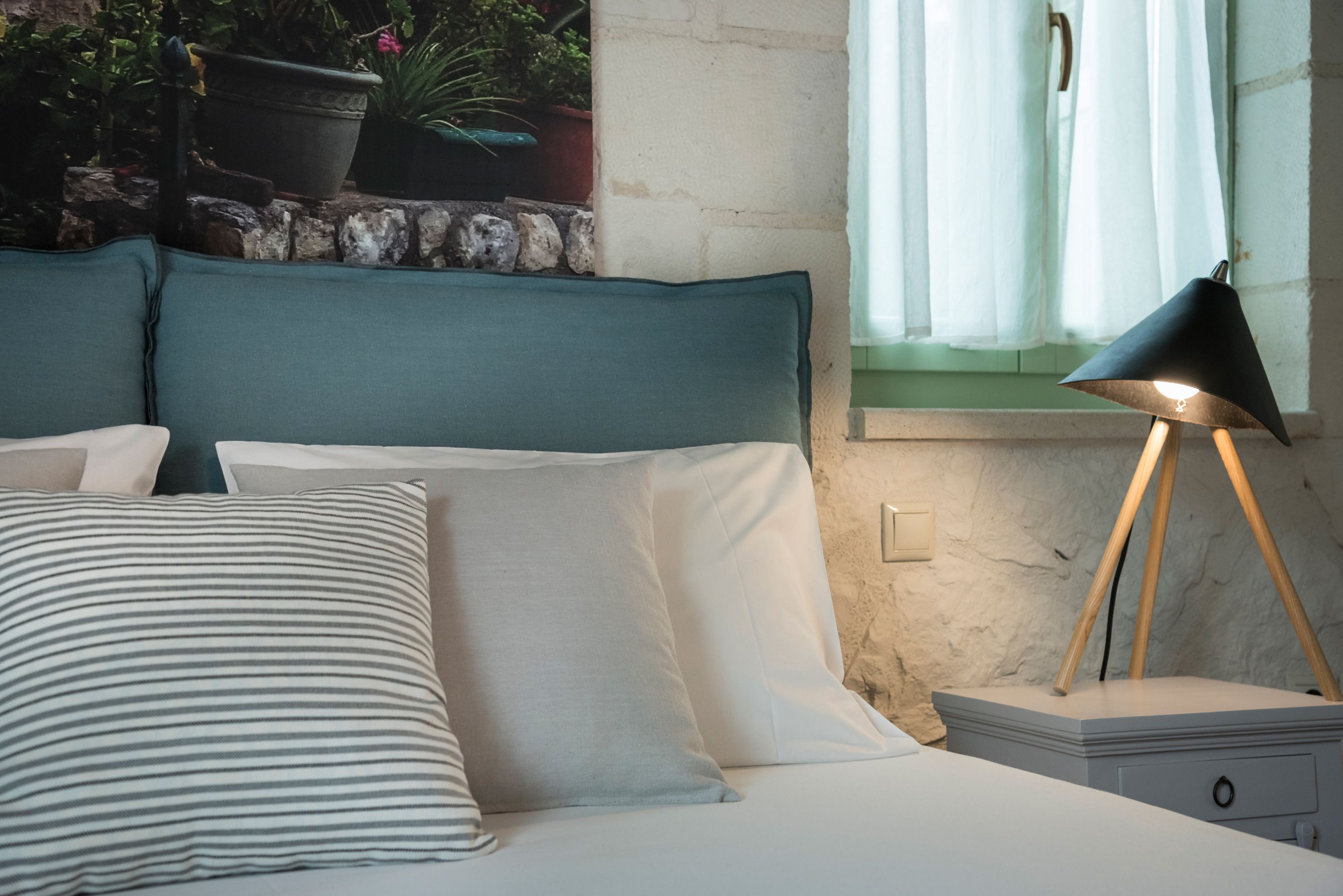 A neatly made bed with striped and solid pillows, a bedside lamp, and a view of a stone wall with potted plants.