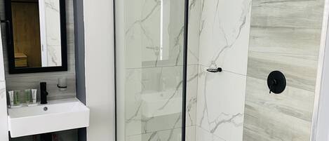 Un baño moderno con ducha de vidrio, un lavabo blanco con artículos de aseo y un estante de madera.