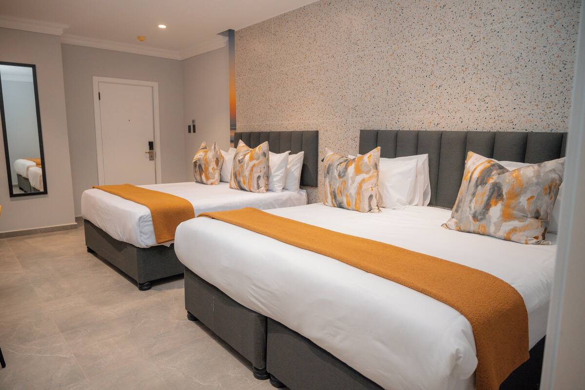A hotel room with two beds, each with a grey headboard, white bedding, and orange throw blankets.