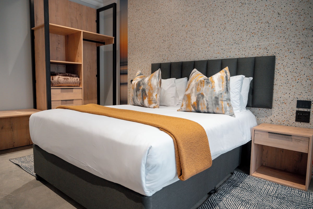 A modern hotel room with a large bed, a wooden wardrobe, and a small bedside table.