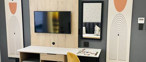 Quarto moderno com televisão de tela plana presa a um painel de madeira, uma escrivaninha pequena com uma cadeira e obras de arte nas paredes.