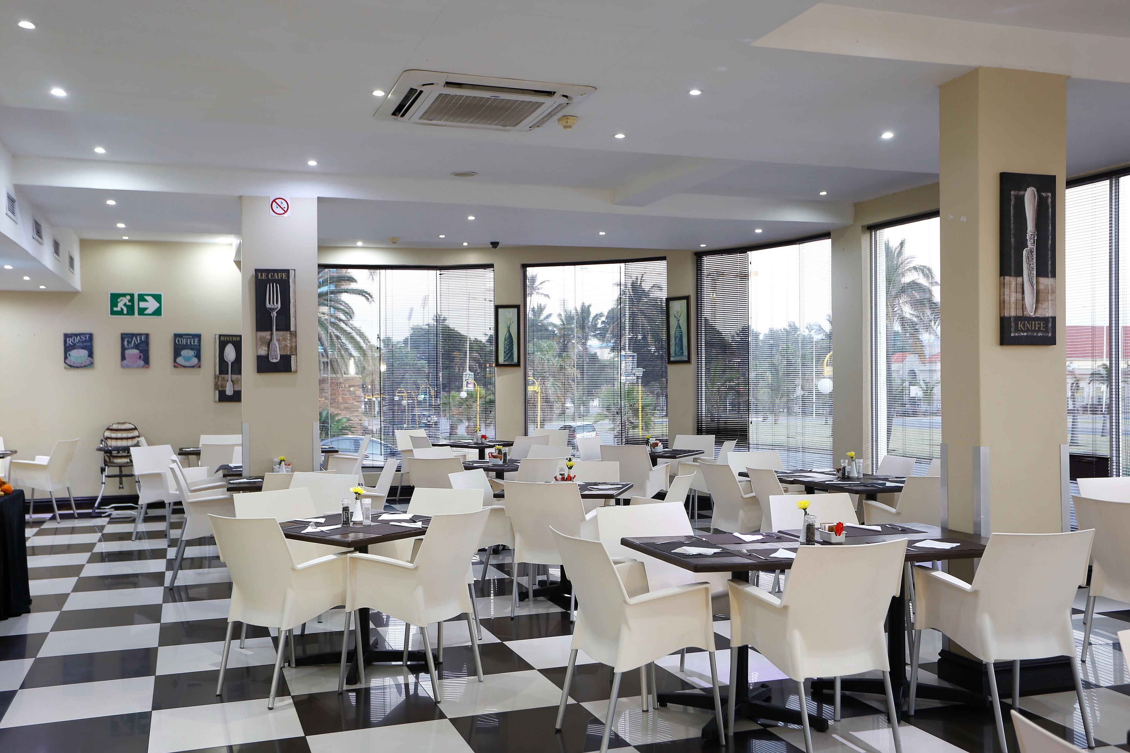 A restaurant with a checkered floor, white chairs, and tables. There are large windows with a view of palm trees and buildings.