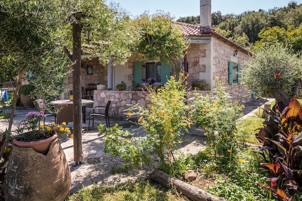 A stone house with a tiled roof, a patio with outdoor furniture, and a garden with various plants and flowers.
